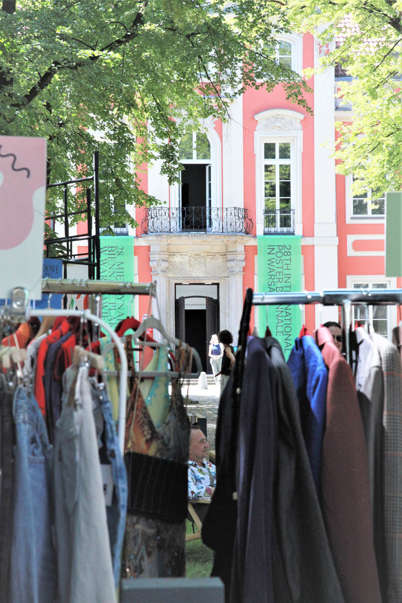Fragment fasady Pałacu Czapskich uchwycony zza stojaków z wiszącymi na nich ubraniami