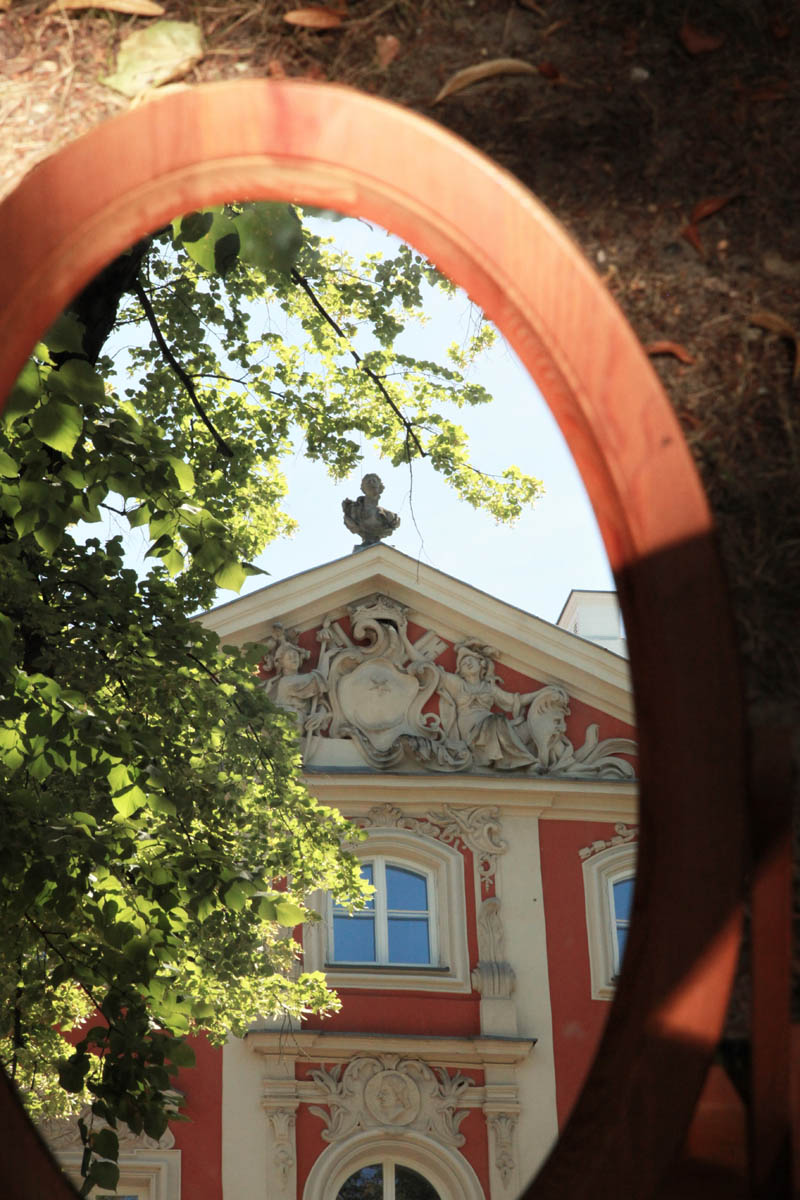 Zdjęcie fragmentu fasady Pałacu Czapskich odbijającej się w owalnym lustrze w drewnianej oprawie