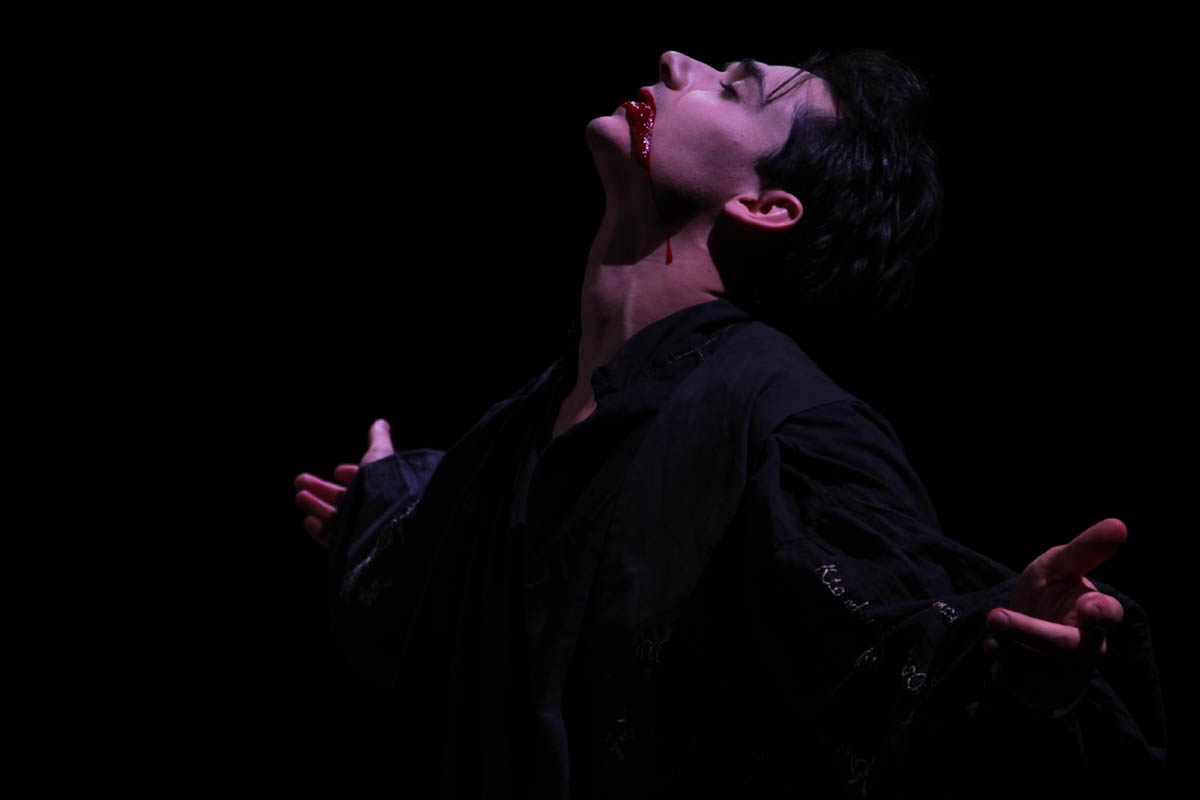 Scena ze spektaklu. Aktor stoi z rozłożonymi rękami i odchyloną głową. Z ust wypływa krew.