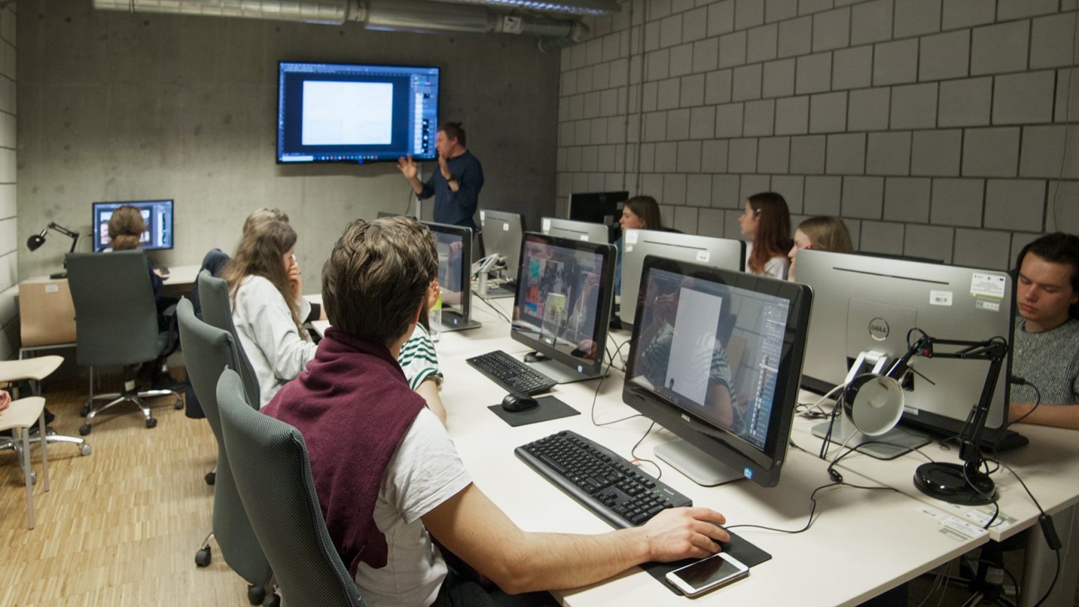 Studenci podczas zajęć w pracowni komputerowej. Prowadzący omawia interfejs programu graficznego wyświetlany na telewizorze zawieszonym na ścianie