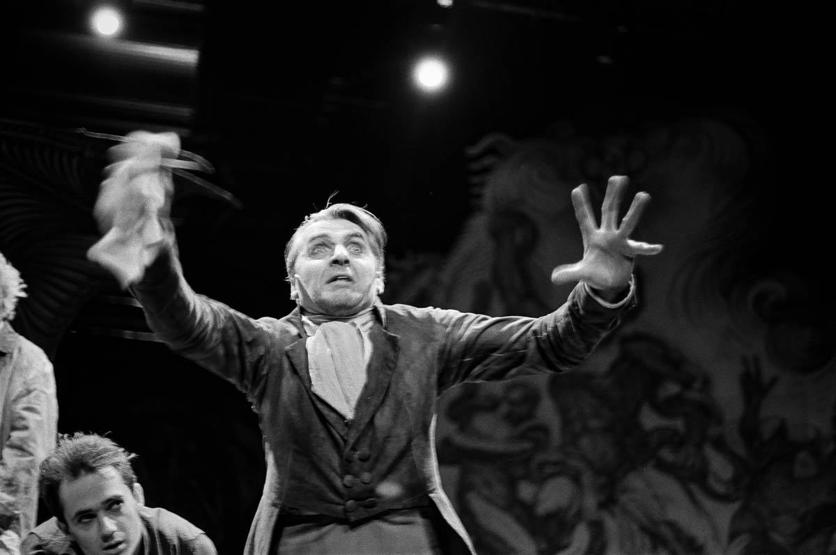 Scena ze spektaklu. Mężczyzna stoi z uniesionymi dłońmi, twarz wyraża emocję