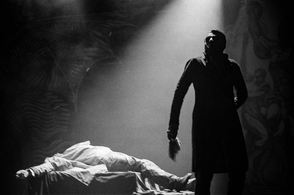 Scena ze spektaklu. Leżaca postać na postumencie, obok stoi mężczyzna w dramatycznej pozie.