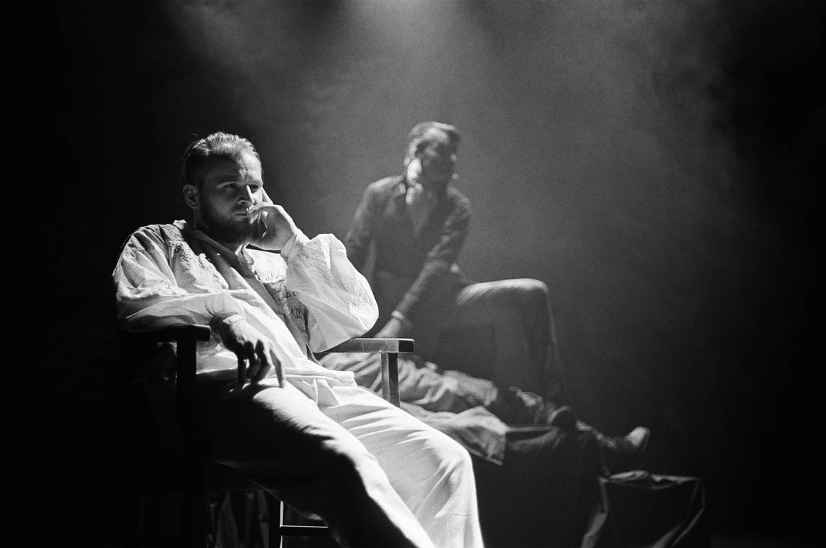 Scena ze spektaklu. Zadumany mężczyzna siedzi w fotelu. Na drugim planie widoczny zarys postaci