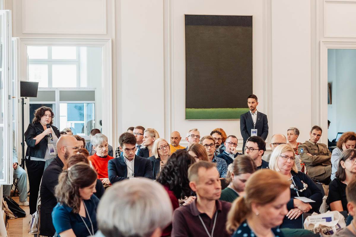 Uczestnicy konferencji siedzą w sali konferencyjnej. Stojąca kobieta trzyma w ręku mikrofon zadając pytanie moderatorowi dyskusji
