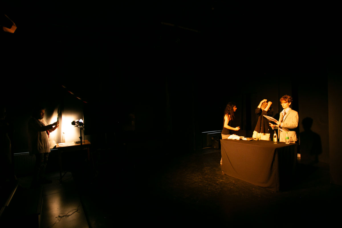 Grupa ludzi na scenie. Mężczyzna i dwie kobiety stoją za stołem pokrytym czarnym suknem. Po lewej stronie przy reflektorze stoi drugi mężczyzna