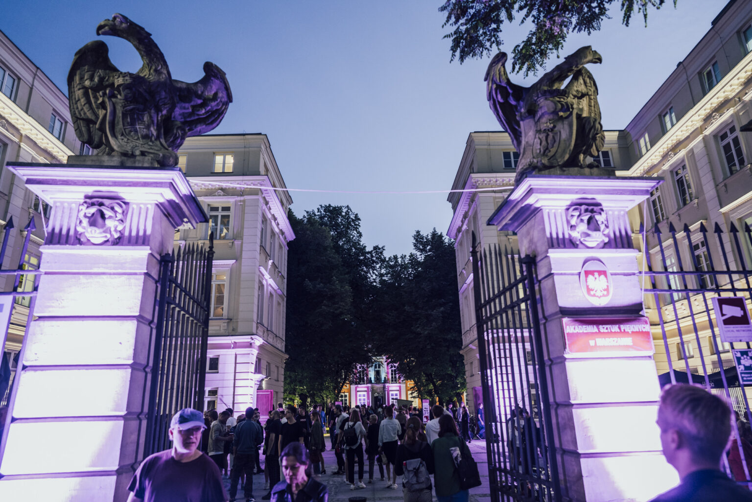 Duża grupa ludzi na dziedzińcu kampusu widoczna przez podświetloną bramę