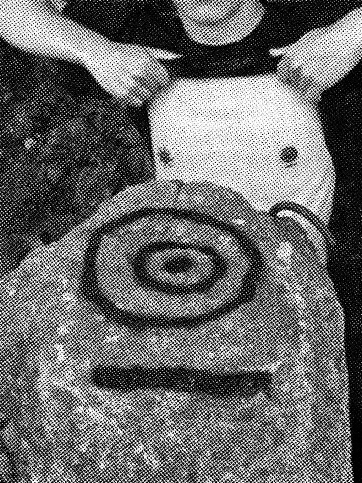 Na pierwszym planie kamień, z narysowanym na nim symbolem za kamieniem stoi mężczyzna podciągający do brody koszulkę