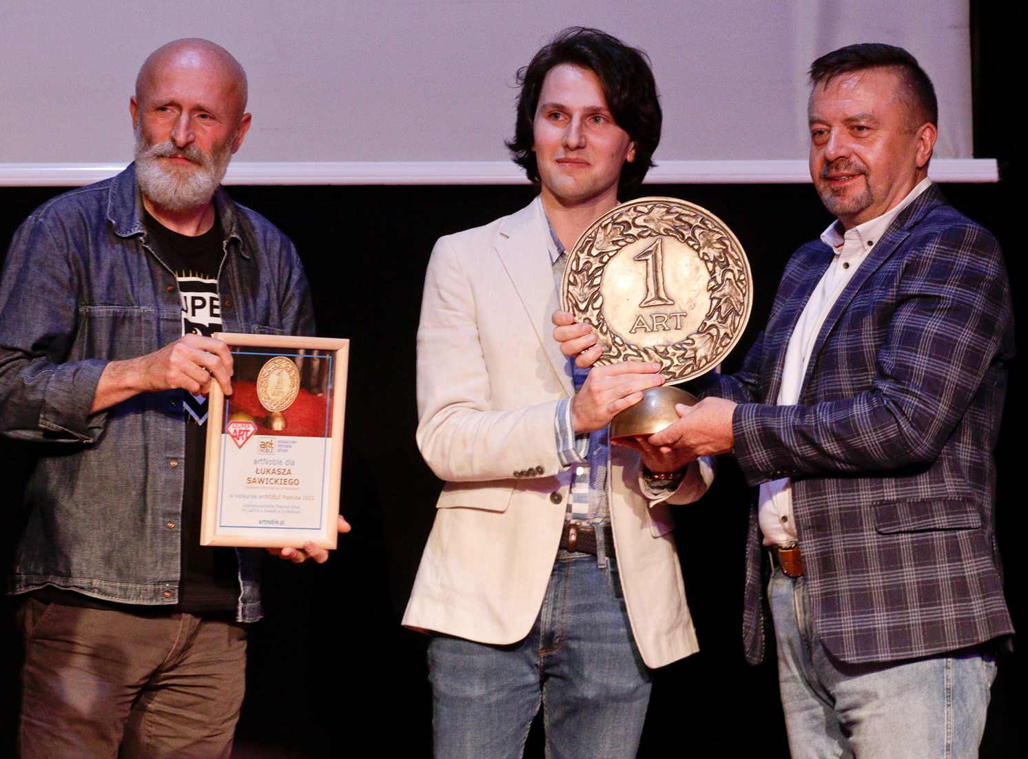Trzy postaci. Po środku mężczyzna ze statuetką.