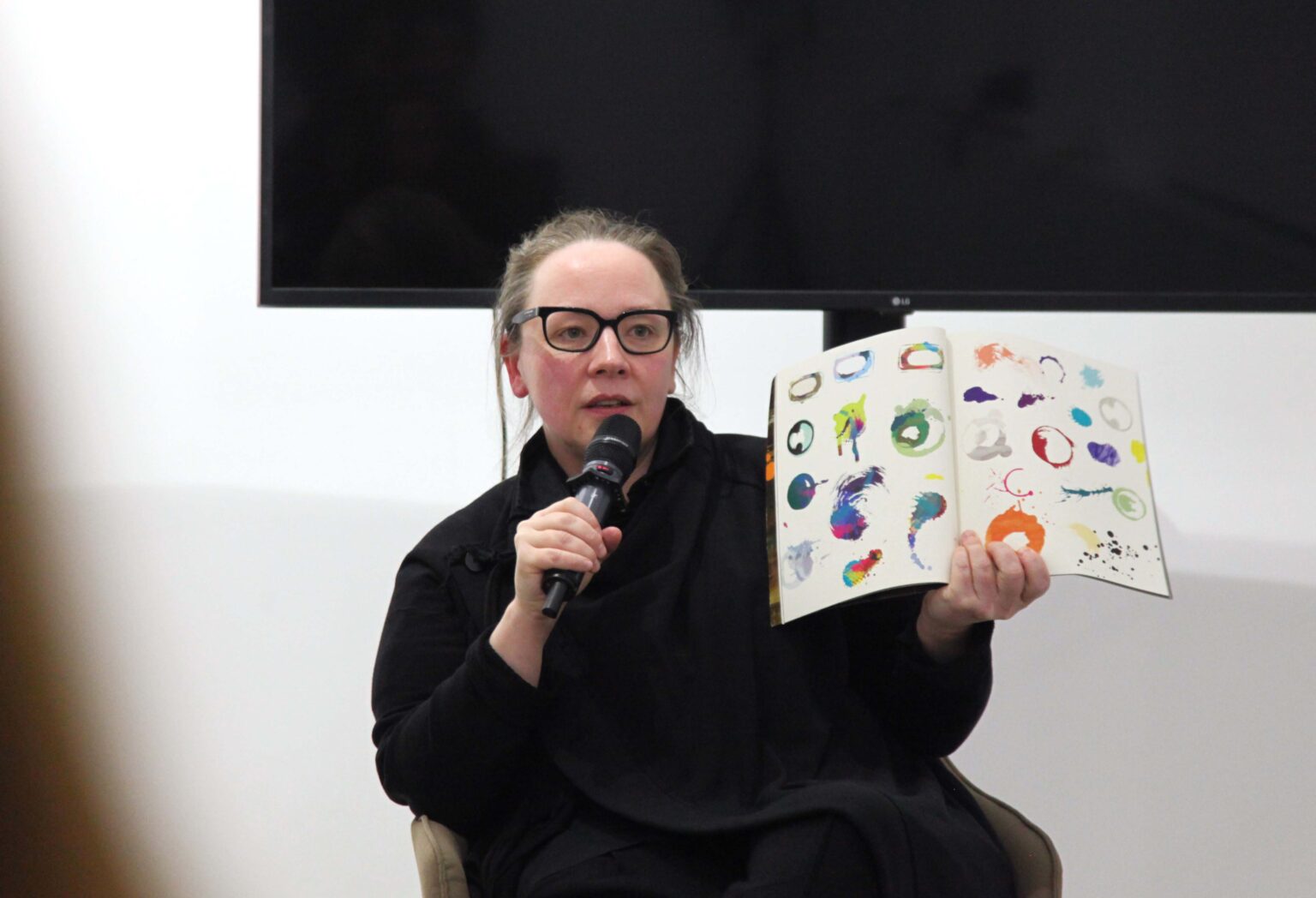 Kobieta mówiąca do mikrofonu. W dłoni trzyma otwartą książkę