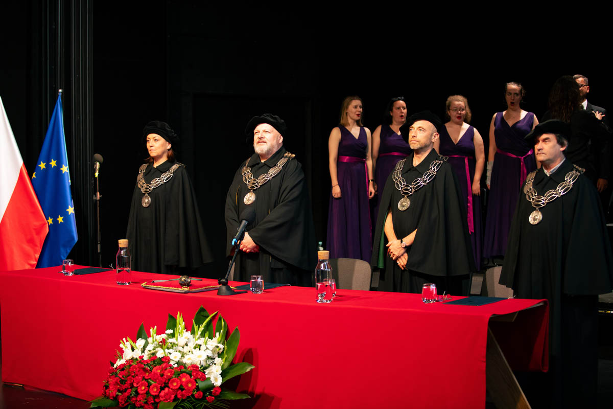 Trzy osoby w uroczystych togach i insygniach akademickich. W tle chór