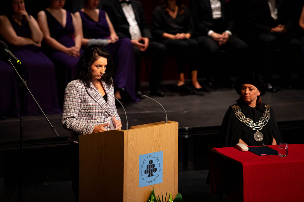 Kobieta na mównicy. Osoby na widowni w tle