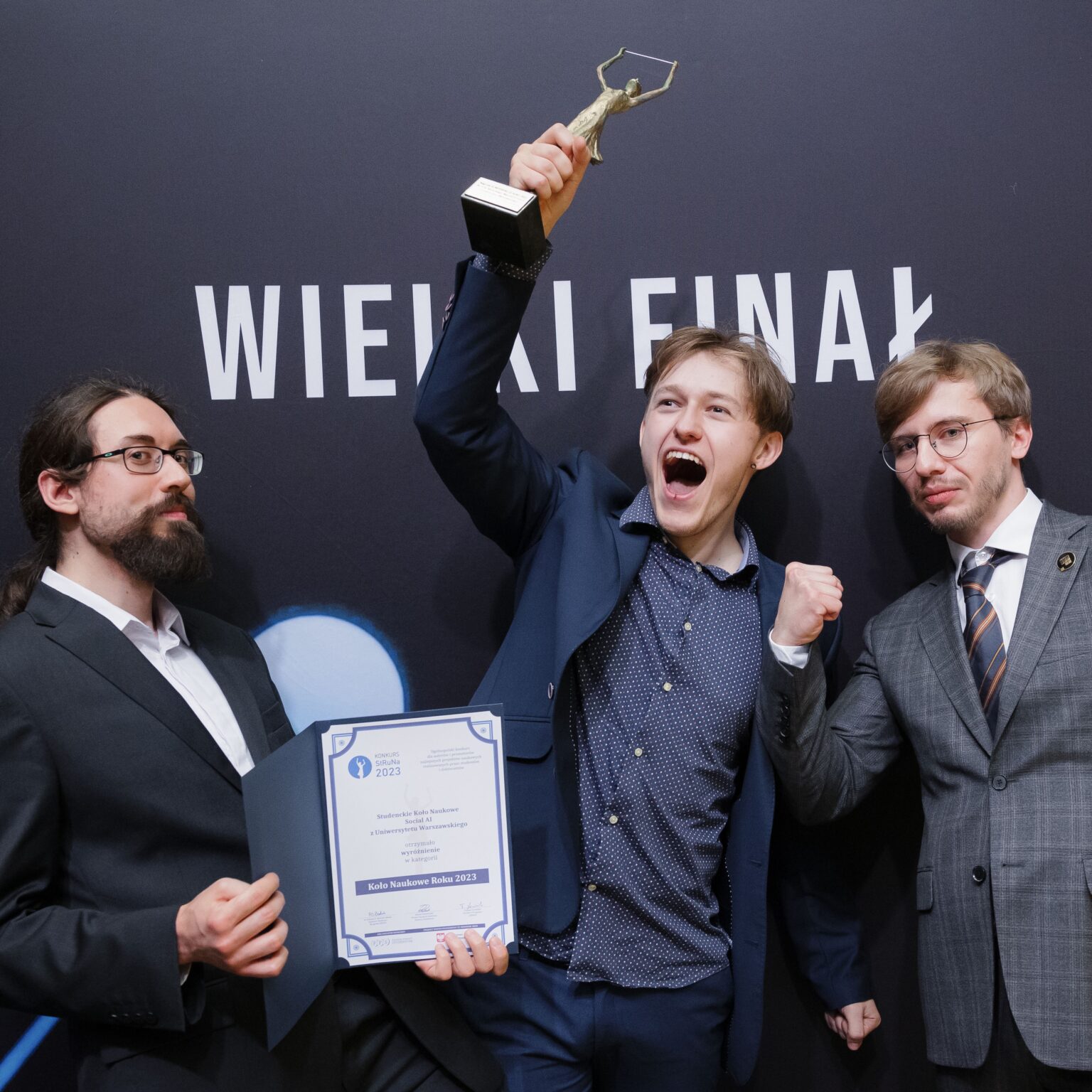 Trzej cieszący się mężczyźni. Jeden z nich trzyma w uniesionej ręce statuetkę w geście zwycięstwa