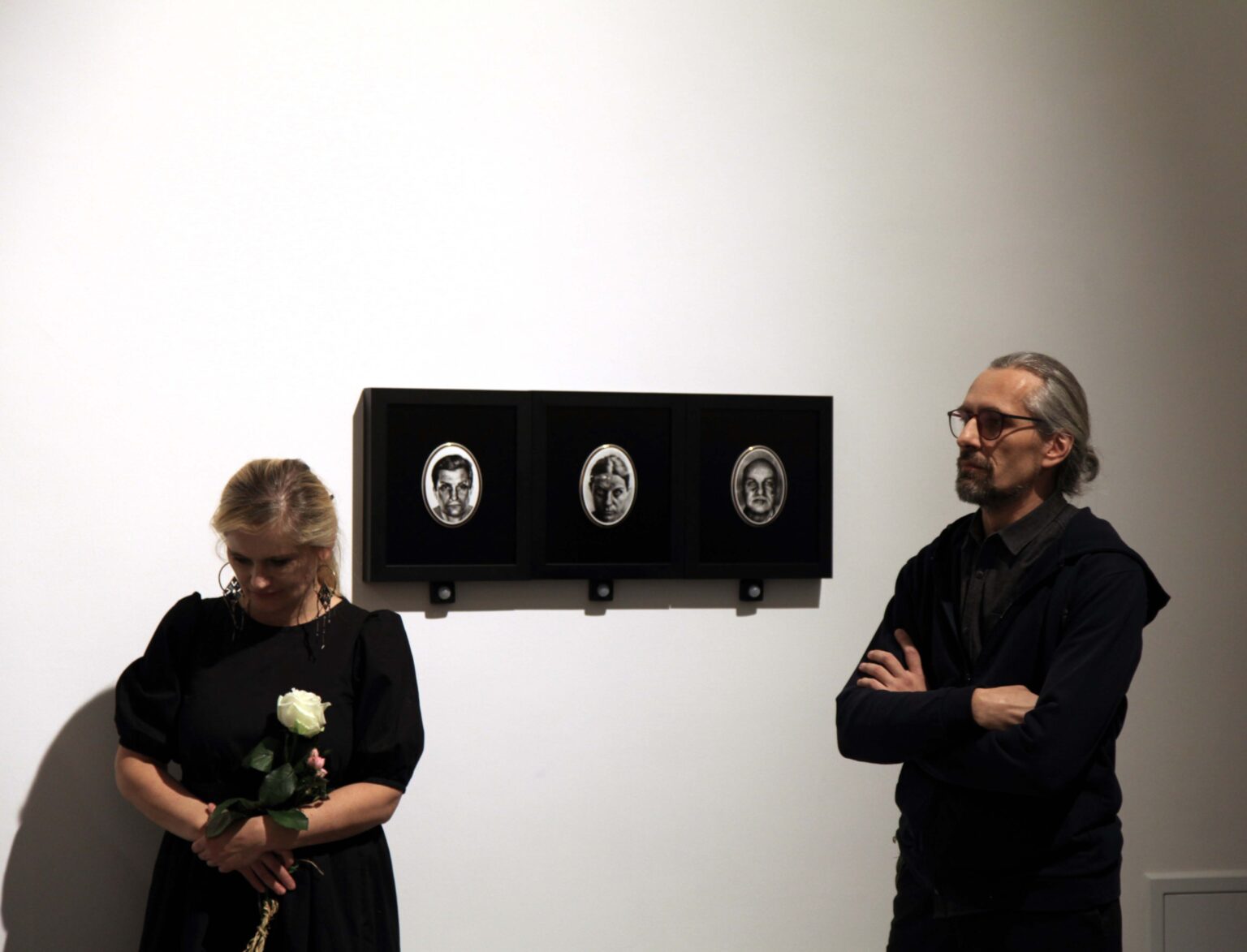 Kobieta z biała różą. Obok mężczyzna. Wystaw w tle.