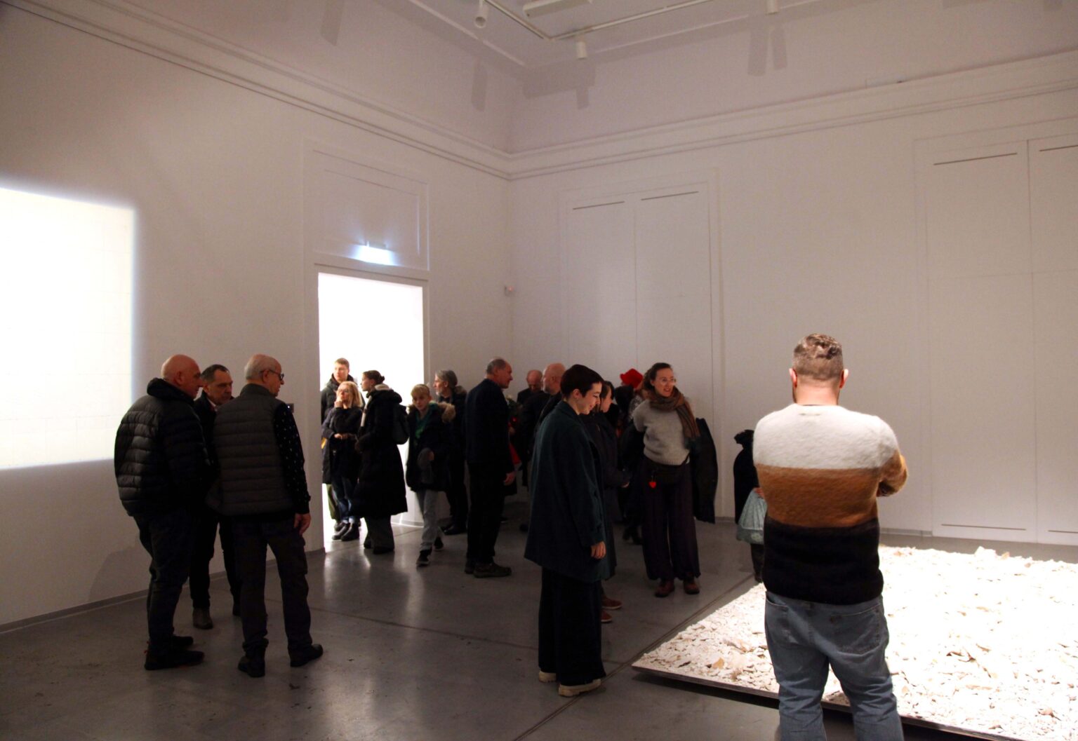 Duża grupa osób w przestrzeni galerii. Eksponat z wysypanych białych kamieni na podłodze