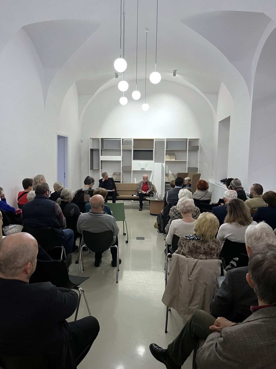 Grupa osób siedzących na krzesłach. W oddali trzy osoby w fotelach