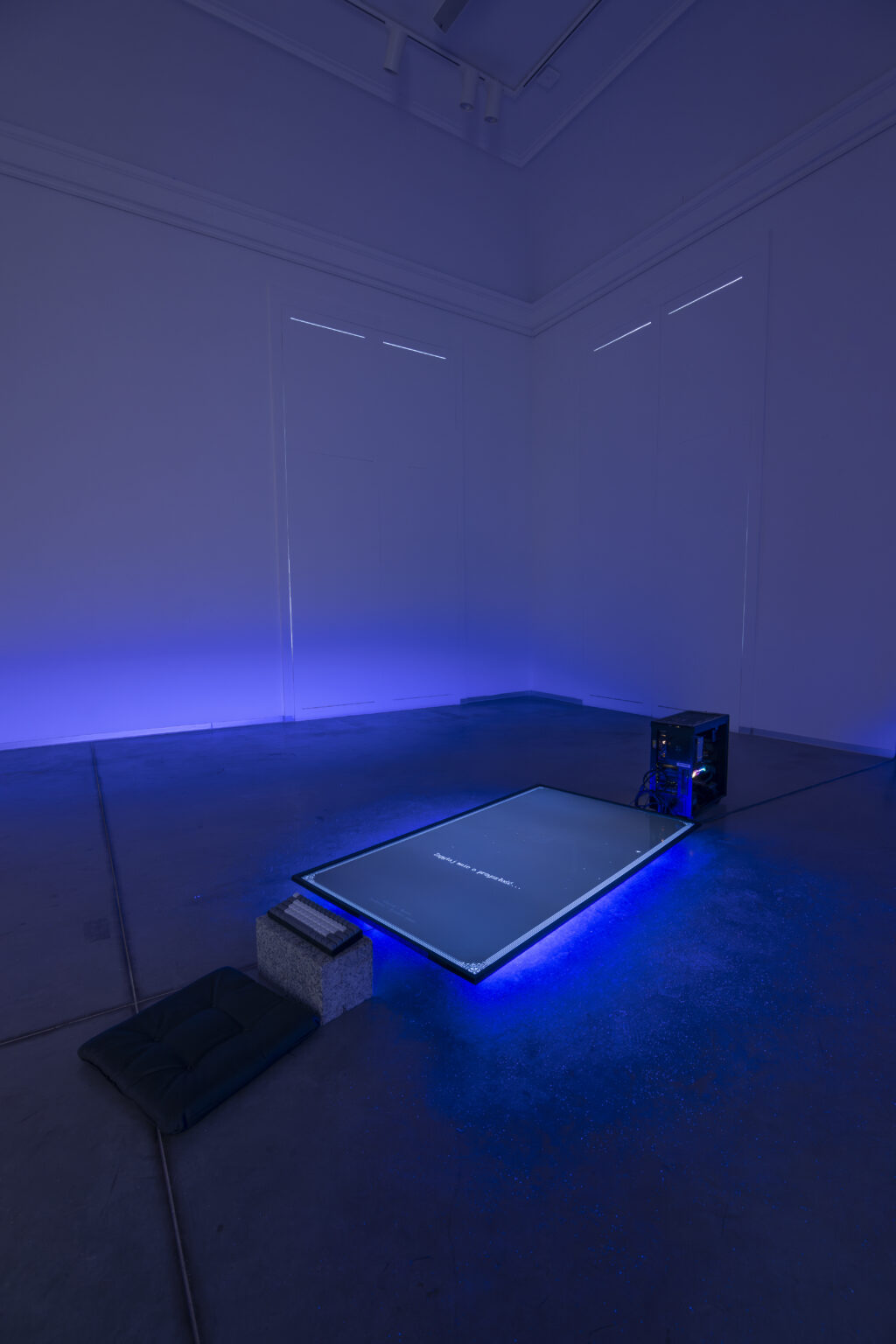 W wyciemnionej przestrzeni na ziemi lezy duży płaski ekranem. Spod ekranu świeci niebieskie światło