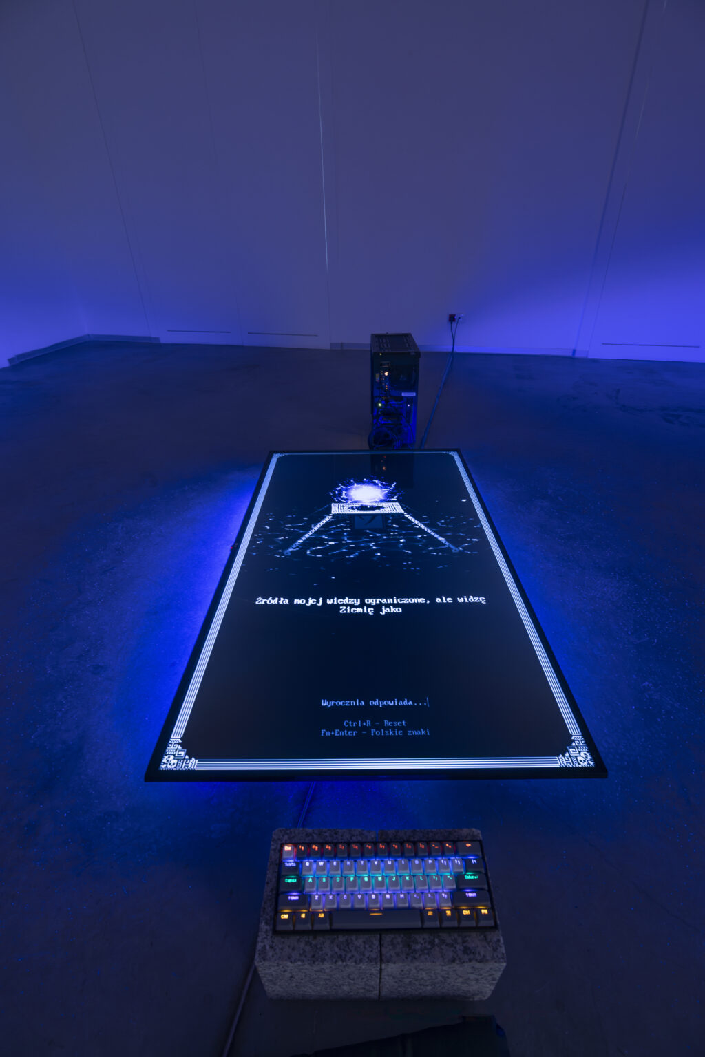 Duży ekran leży na podłodze, spod niego świeci niebieskie światło. Na ekranie tekst wygenerowany przez sztuczną inteligencję