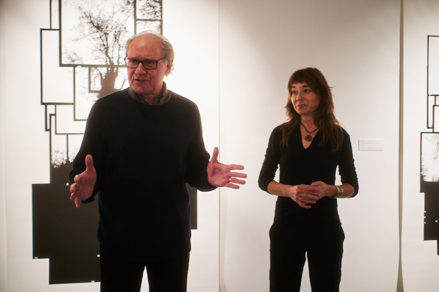 Dwie osoby - mężczyzna i kobieta ubrani na ciemno. Są w trakcie przemowy na otwarciu wystawy