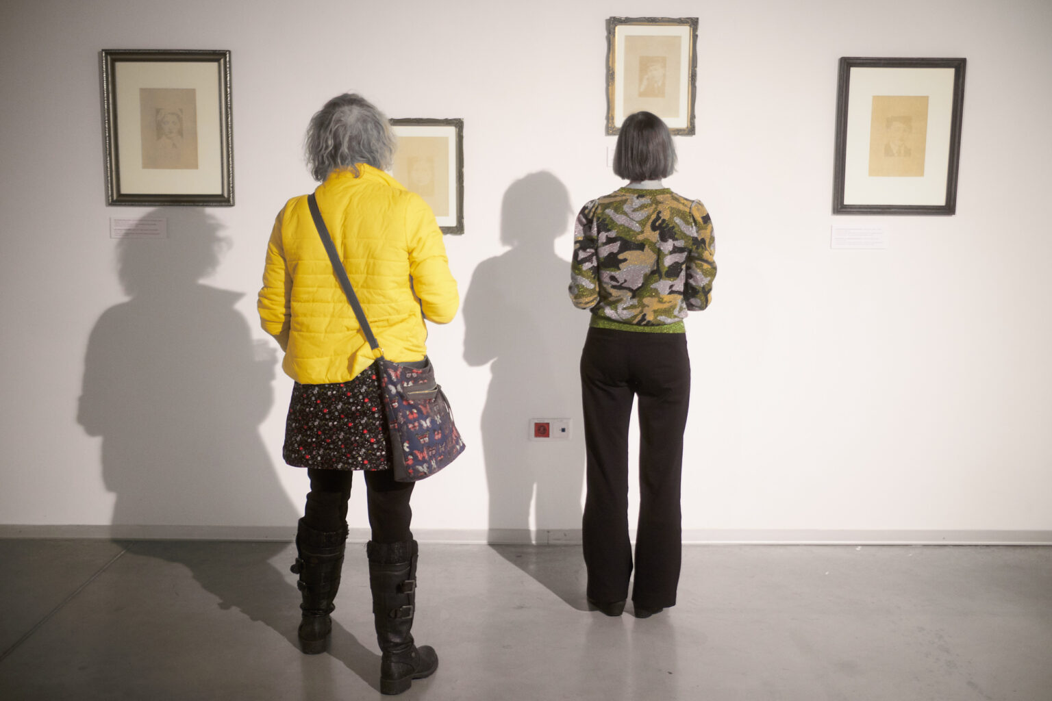 Widok wystawy. Dwie kobiety stoją tyłem oglądając wiszące na ścianie oprawione w ramki grafiki