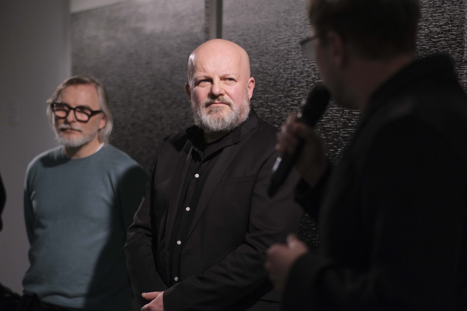 Trzech mężczyzn. Jeden z nich, zwrócony tyłem, mówi do mikrofonu