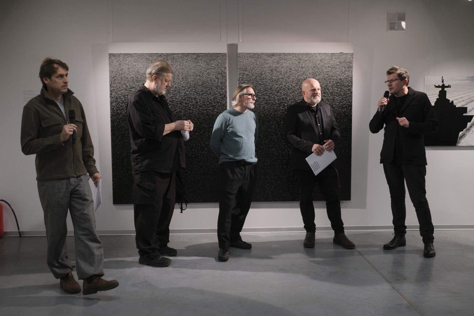 Pięciu mężczyzn stoi na tle wystawy. Pierwszy po prawej mówi do mikrofonu, który trzyma w dłoni.