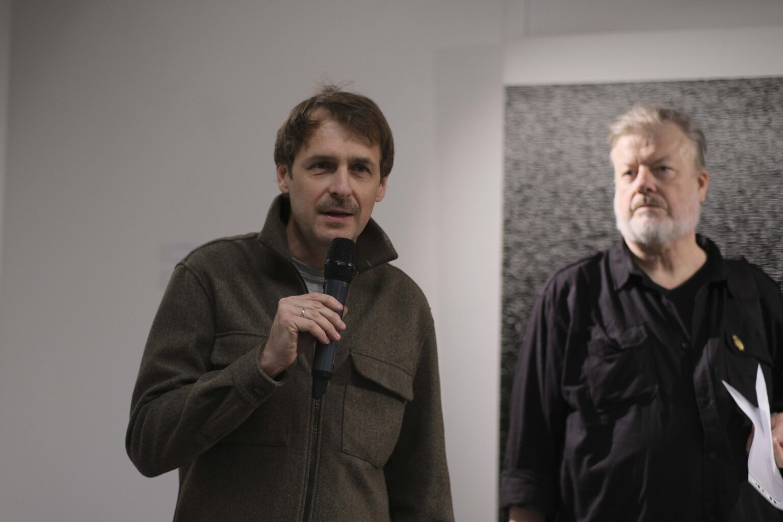 Dwóch mężczyzn na tle obrazu. Stojacy po lewej mówi do mikrofonu