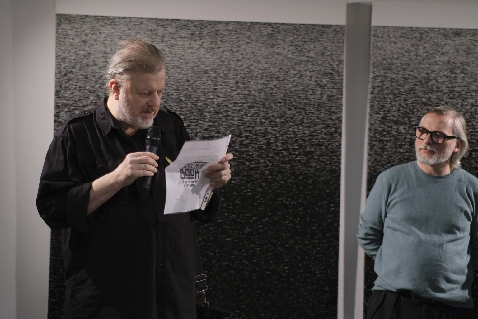 Mężczyzna przemawia. W ręku trzyma kartkę papieru. Monochromatyczny obraz w tle