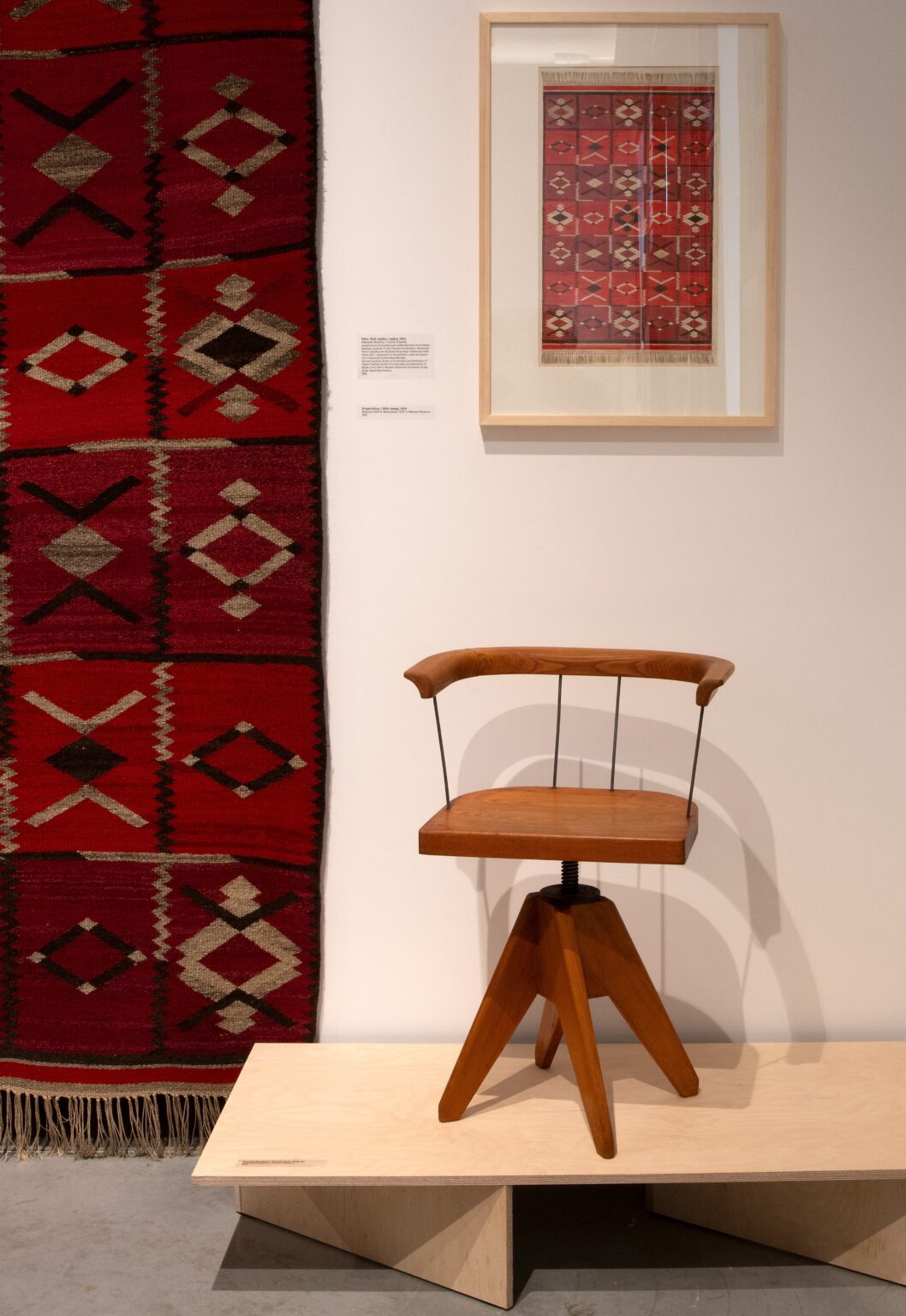Widok wystawy. Od lewej: czerwona wzorzysta tkanina na ścianie, drewniane krzesło obrotowe na podeście; powyżej niego zdjęcie tkaniny oprawione w ramę