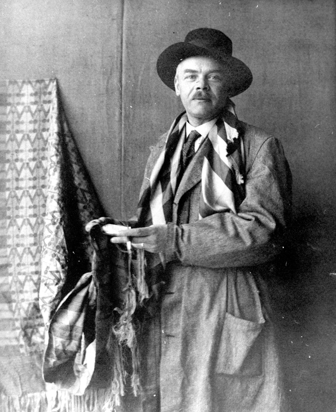 Czarni-biały portret mężczyzny w prochowcu i kapeluszu. Z rękach trzyma fragment tkaniny rozwieszonej obok.