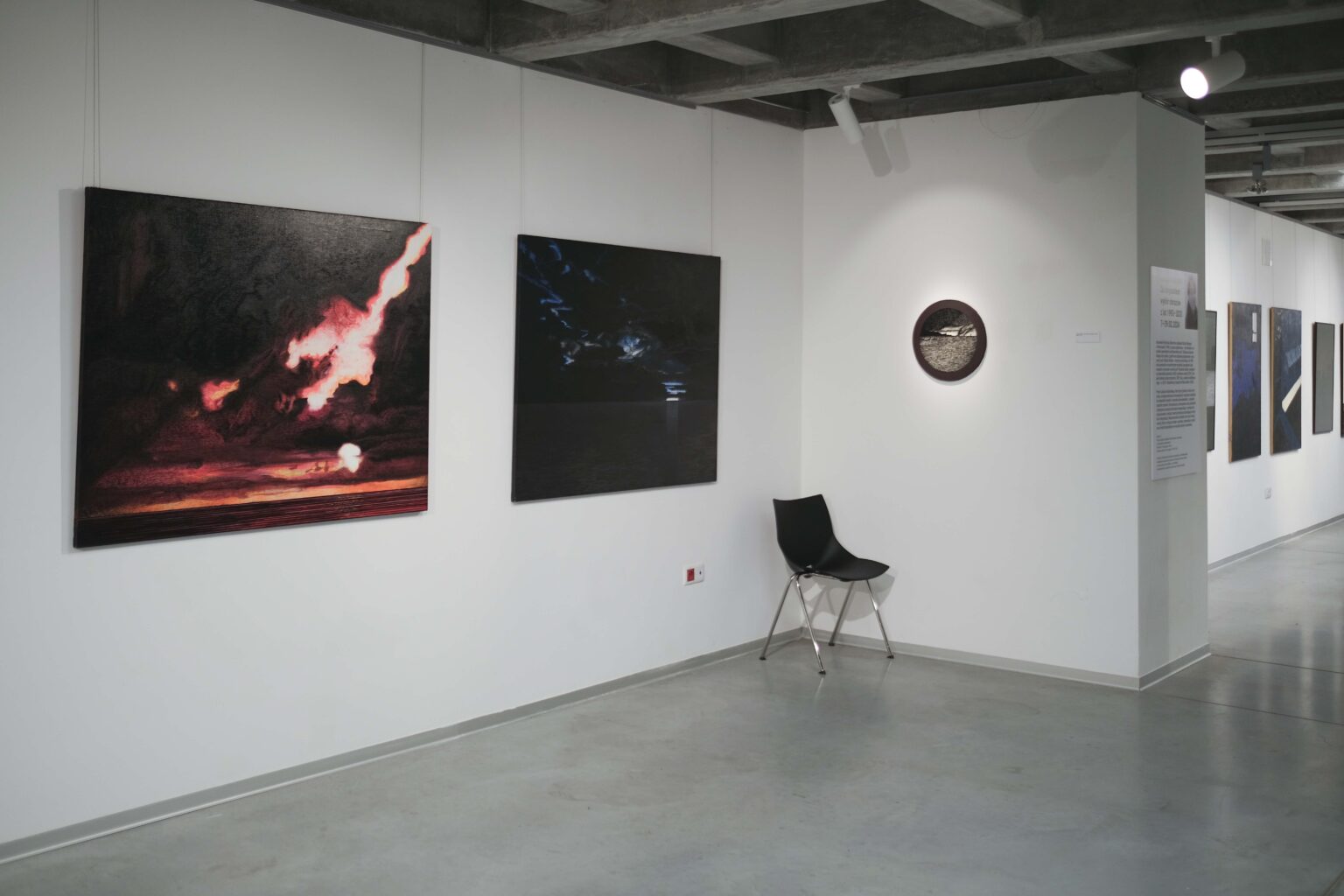 Widok wystawy. Trzy obrazy, jeden z nich w okrągłym kształcie. Krzesło w roku sali