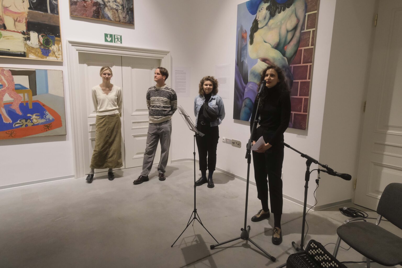 Cztery osoby stoją obok siebie. Kobieta po prawej przy mikrofonie. Obrazy w tle