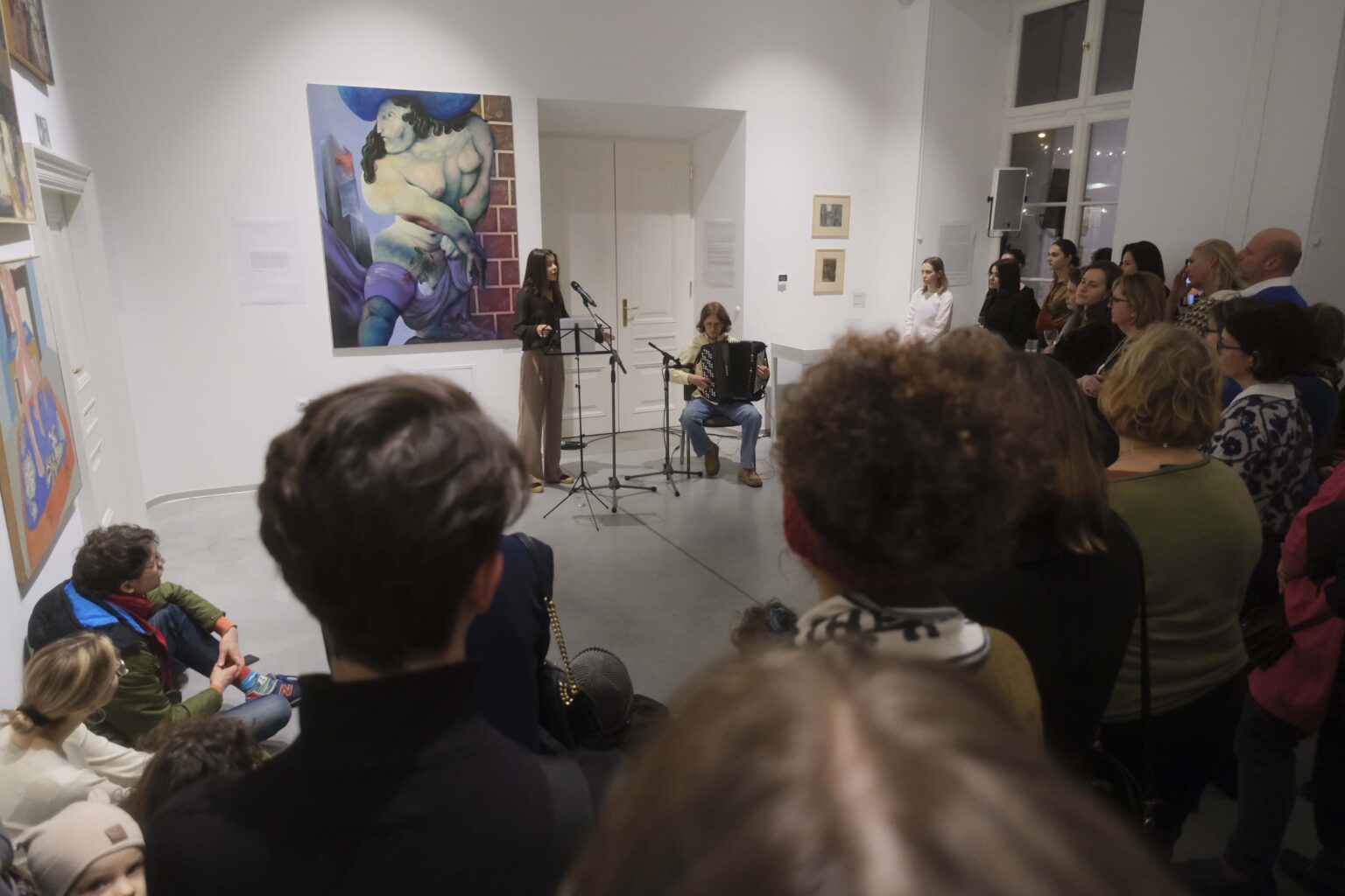 Zwiedzający słuchają koncertu. Przed nimi kobieta stoi przy mikrofonie, mężczyzna gra na akordeonie