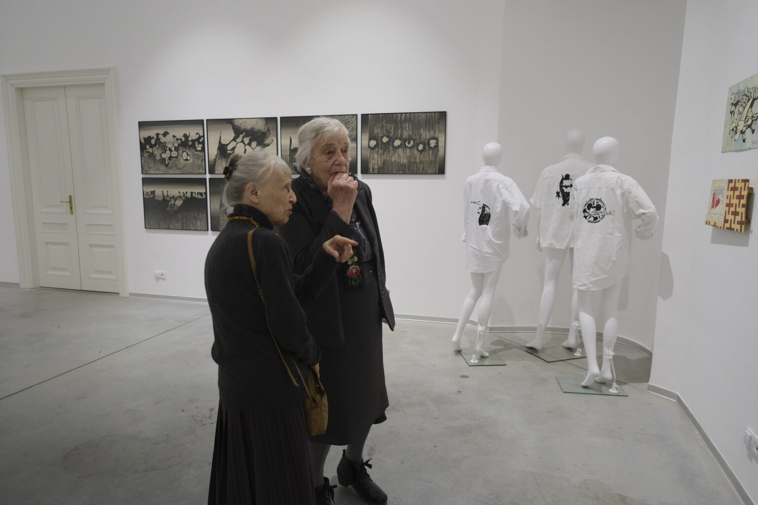 Dwie kobiety oglądają ekspozycję. W prawym narożniku trzy ubrane na biało manekiny odwrócone tyłem