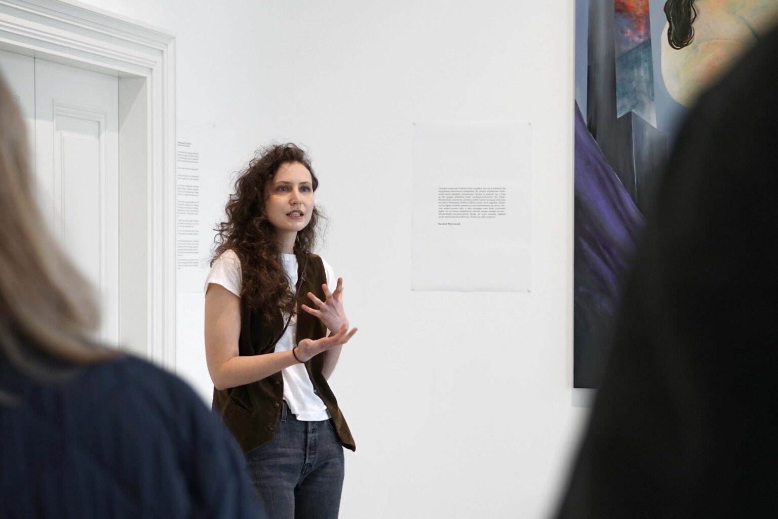 Długowłosa kobieta opowiada gestykulując, w tle wystawa