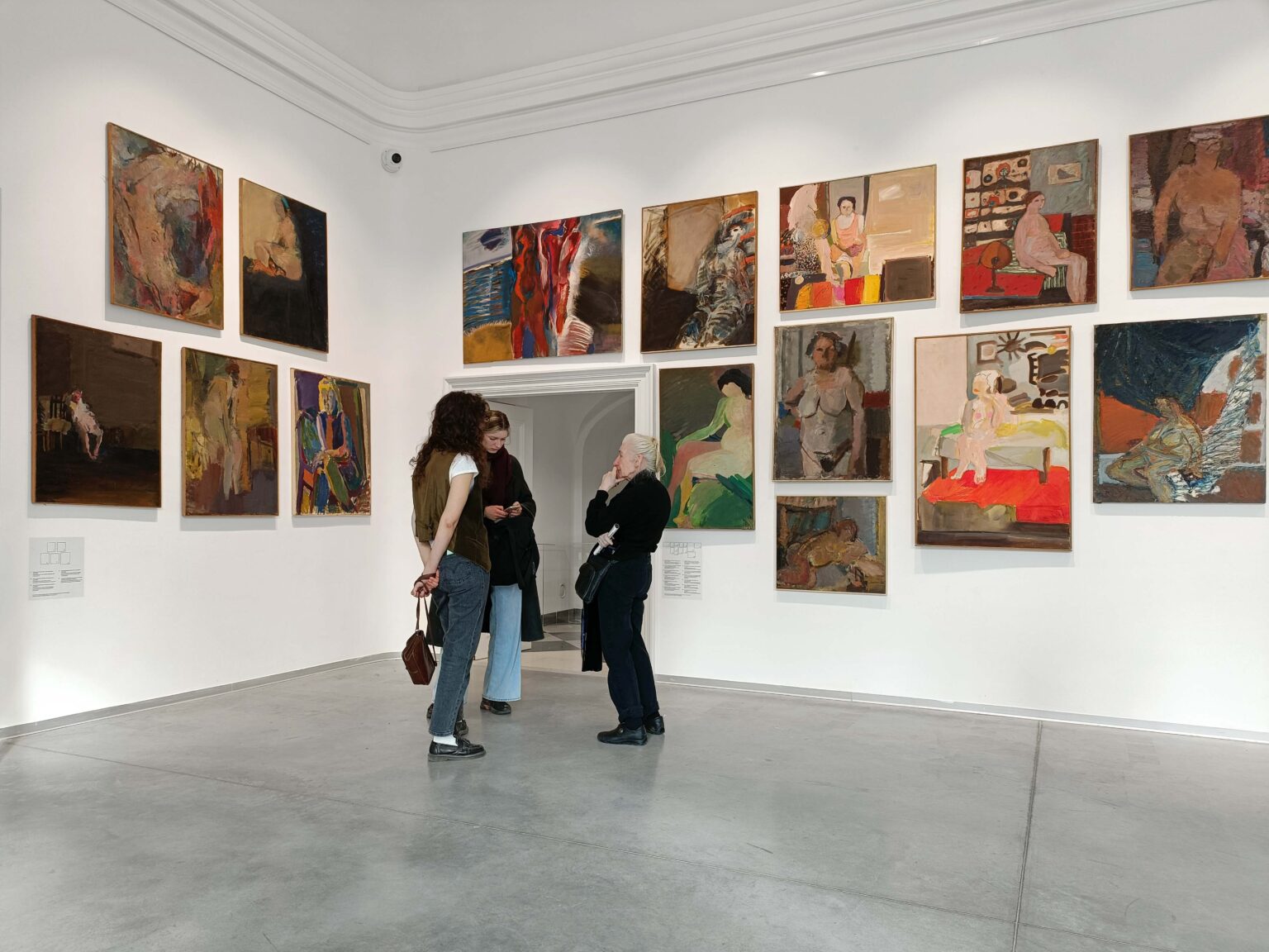 Trzy osoby w przestrzeni galerii. Dużo barwnych obrazów na ścianach