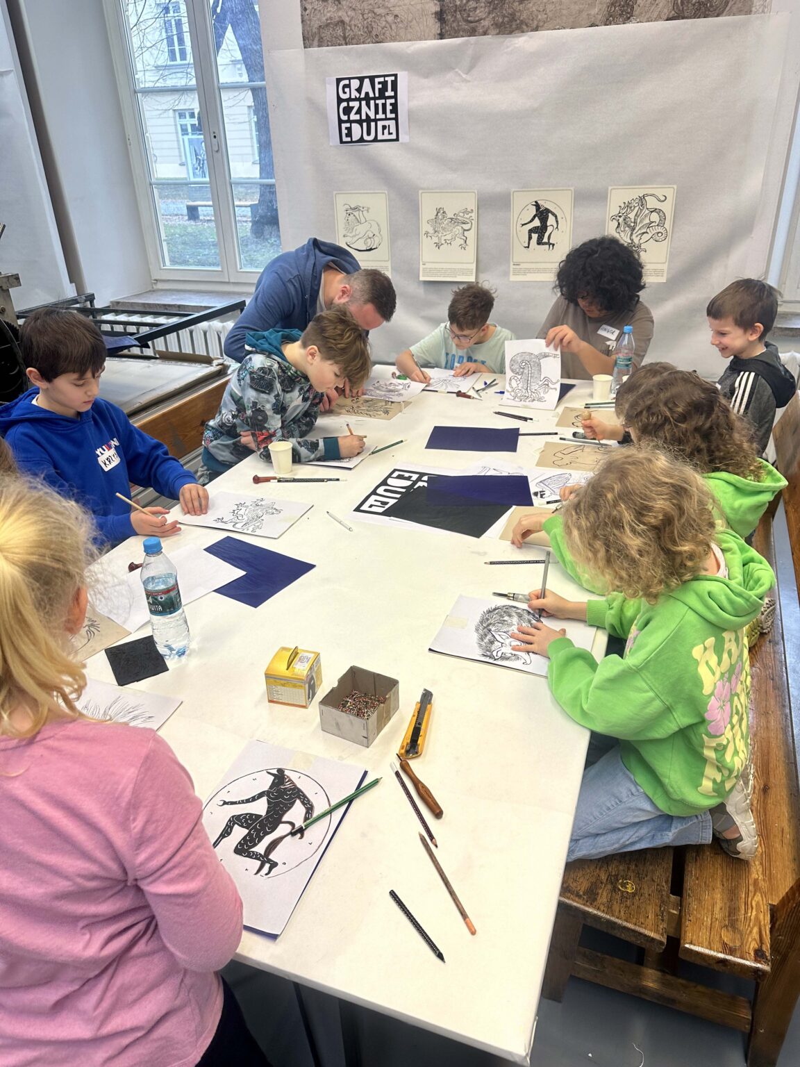 Grupa dzieci przy dużym stole podczas warsztatów. Na stole przybory do rysowania