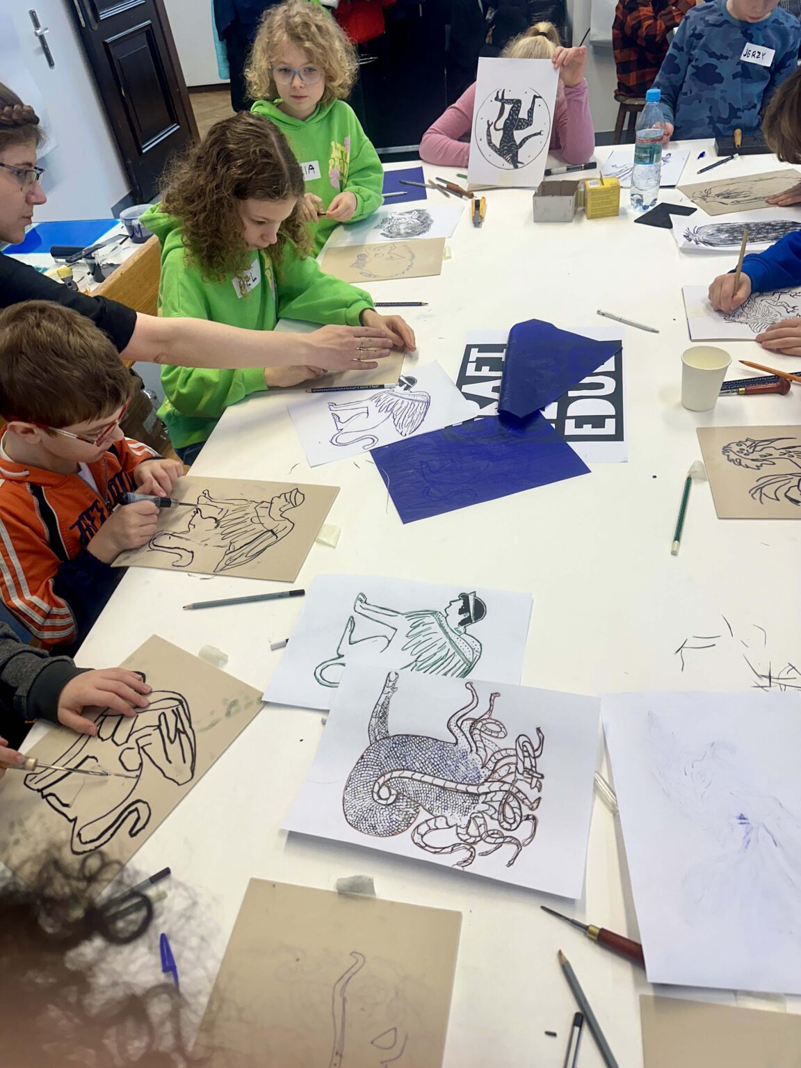 Grupa dzieci przy dużym stole podczas warsztatów. Na stole przybory do rysowania