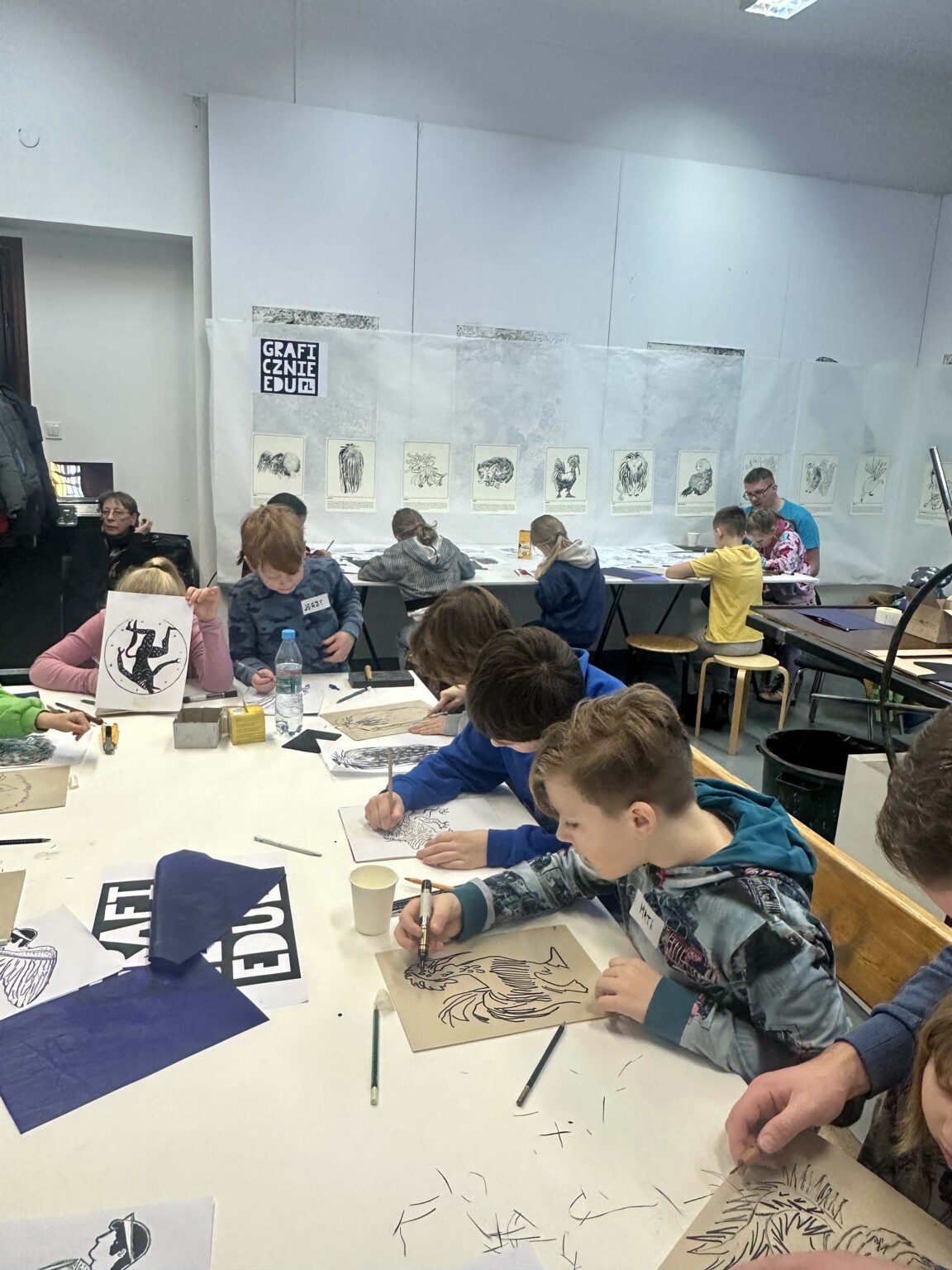 Grupa dzieci przy dużym stole podczas warsztatów. Na stole przybory do rysowania