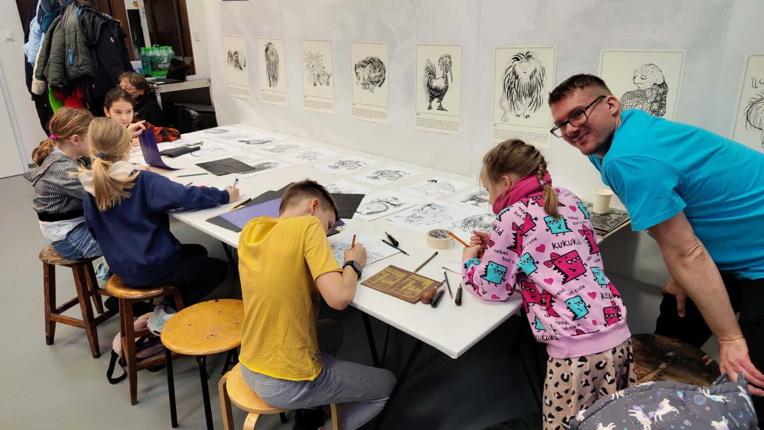 Grupa dzieci przy dużym stole podczas warsztatów. Na stole przybory do rysowania