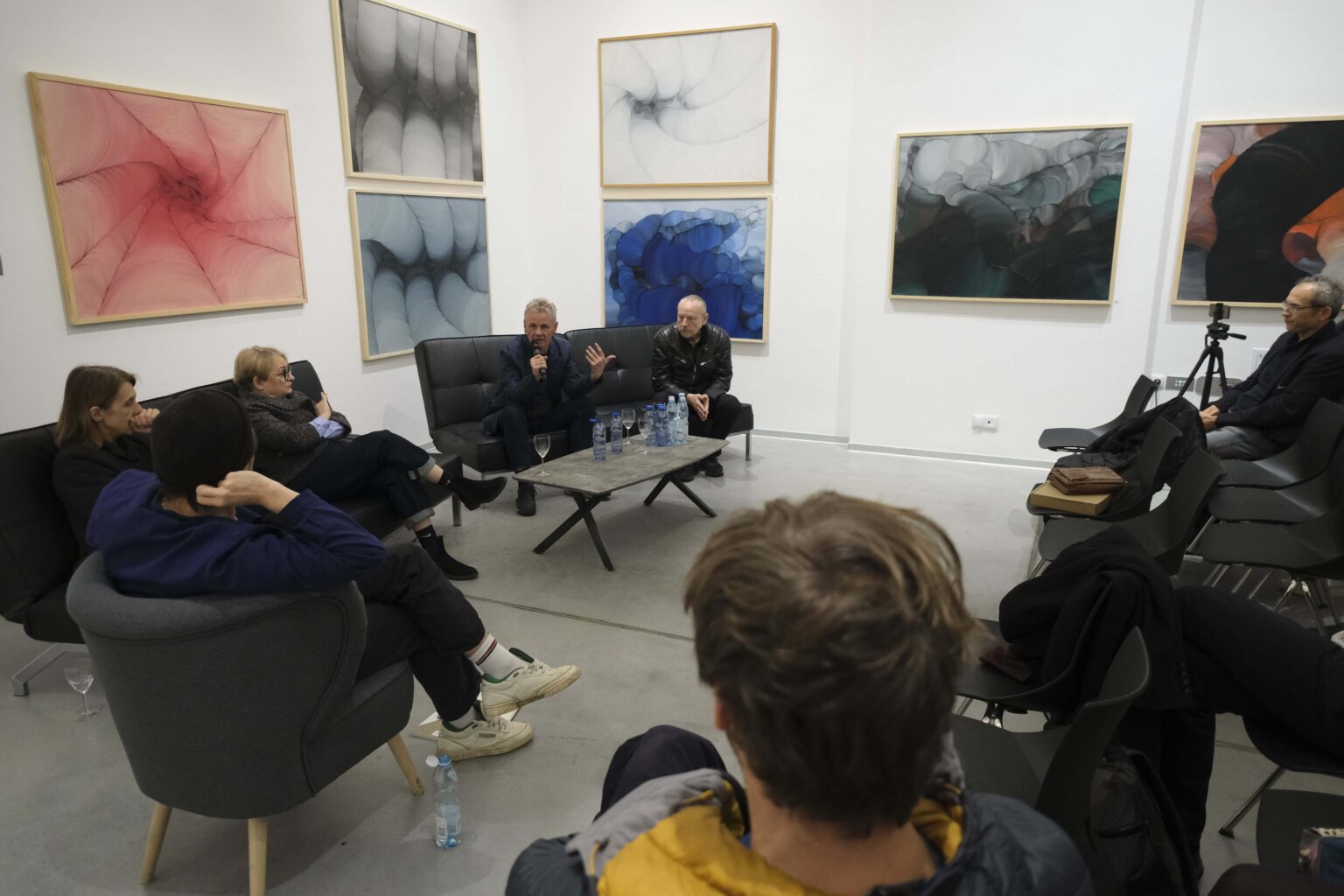 Kilka osób rozmawia na kanapie w przestrzeni galerii. Jedna z nich mówi do mikrofonu. Widać publiczność przysłuchującą się rozmowie
