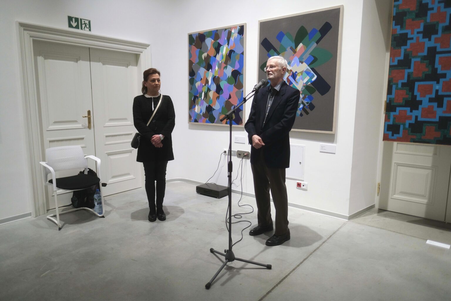 Kobieta, obok mężczyzna przy mikrofonie. Kolorowe obrazy w tle