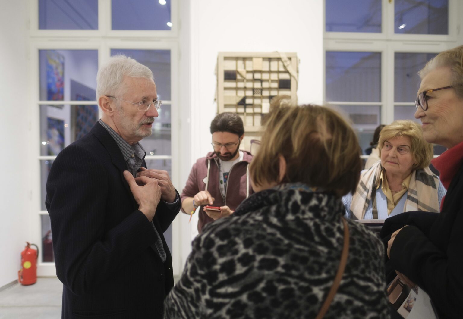 Mężczyzna rozmawia z kilkoma otaczającymi go osobami. Obraz w tle