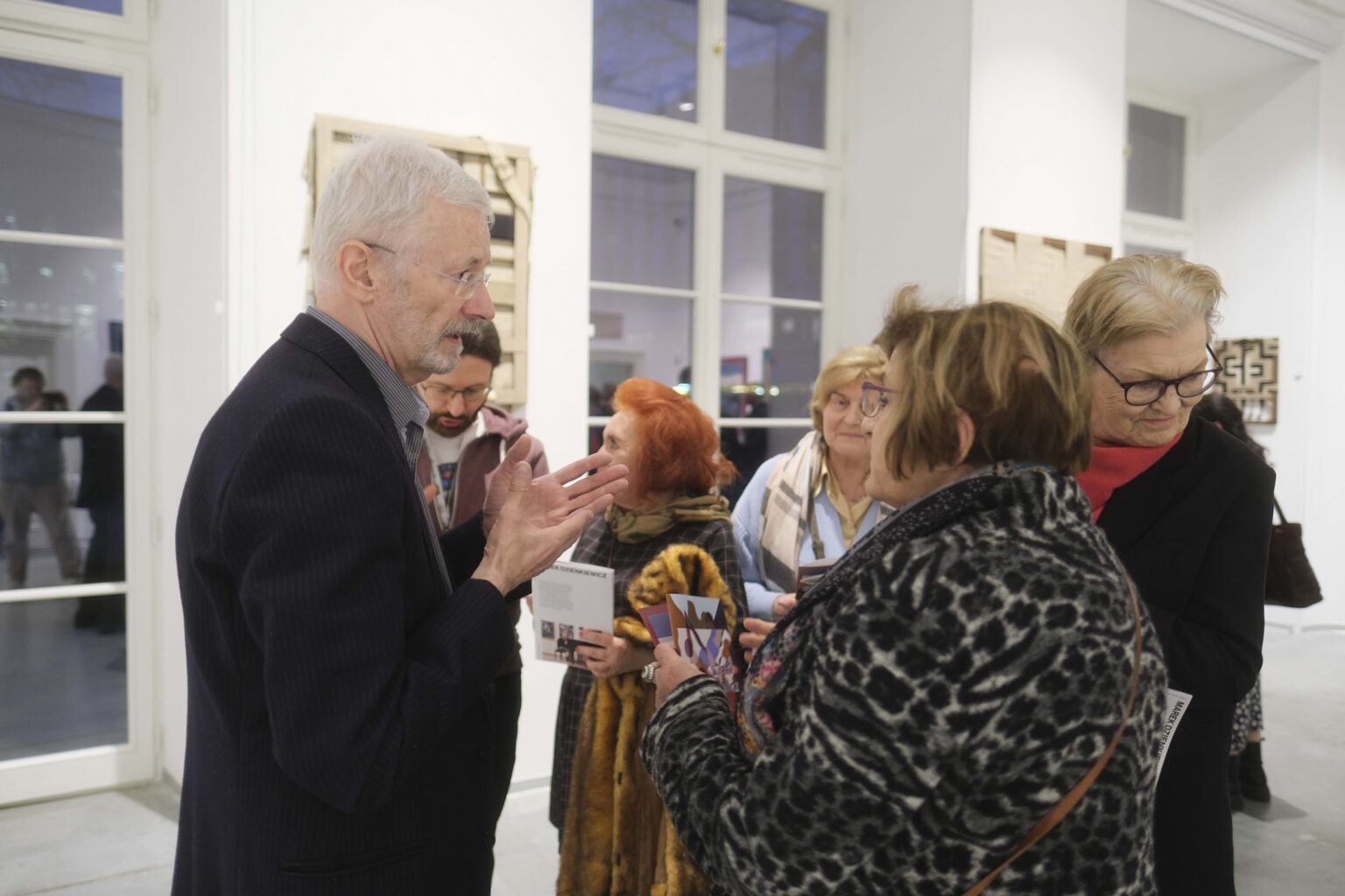 Mężczyzna rozmawia z kilkoma otaczającymi go osobami. Wystawa w tle