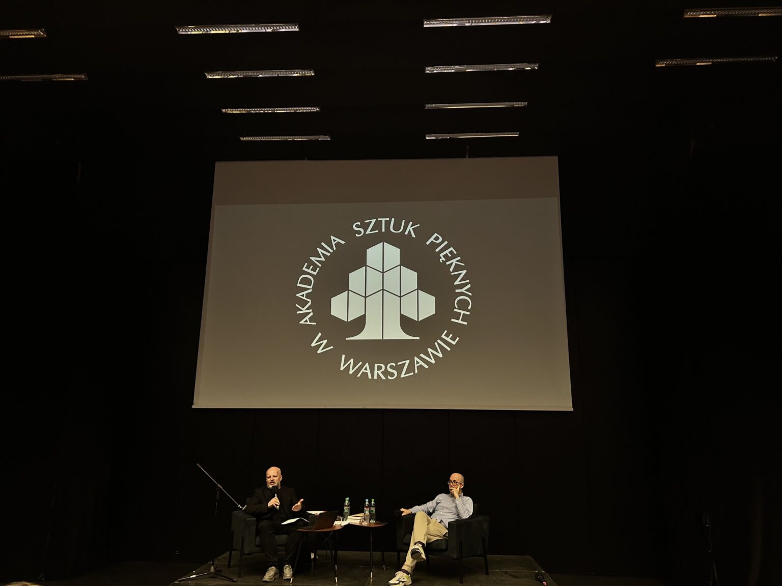 Dwie osoby siedzą i rozmawiją. Nad nimi widnieje logo ASP w Warszawie wyświetlane na ekranie