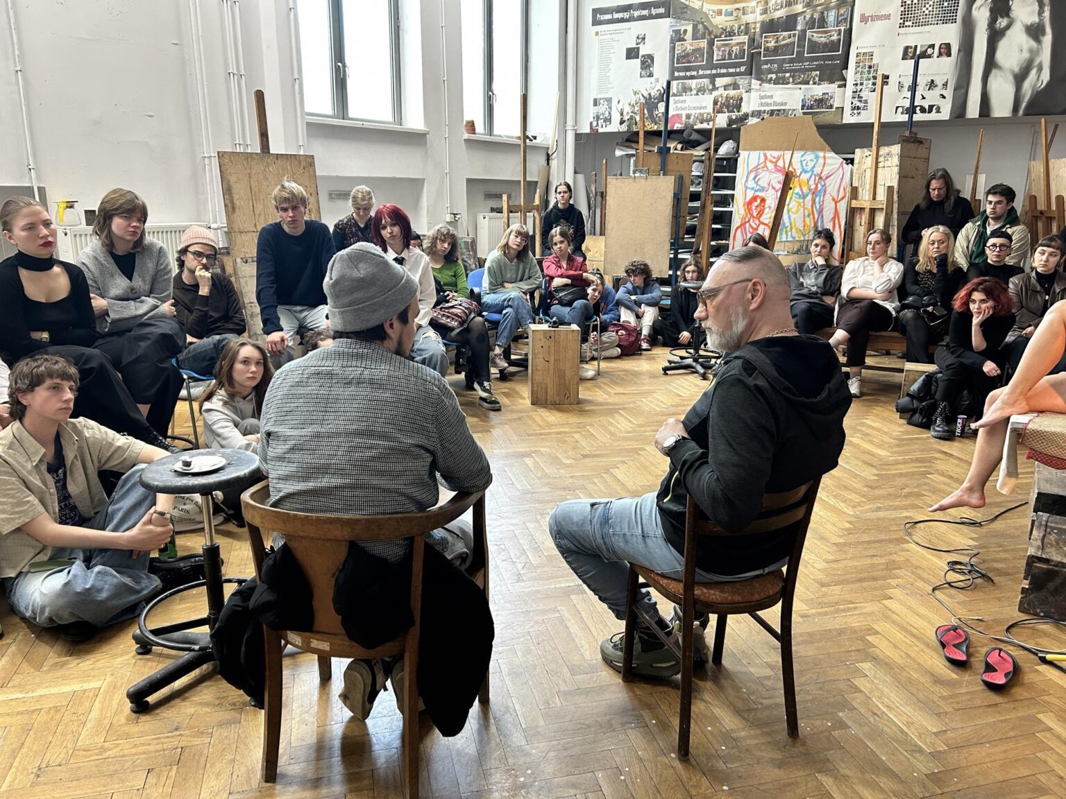 Dwóch mężczyzn tyłem siedzi na krzesłach. Przed nimi grupa osób na krzesłach