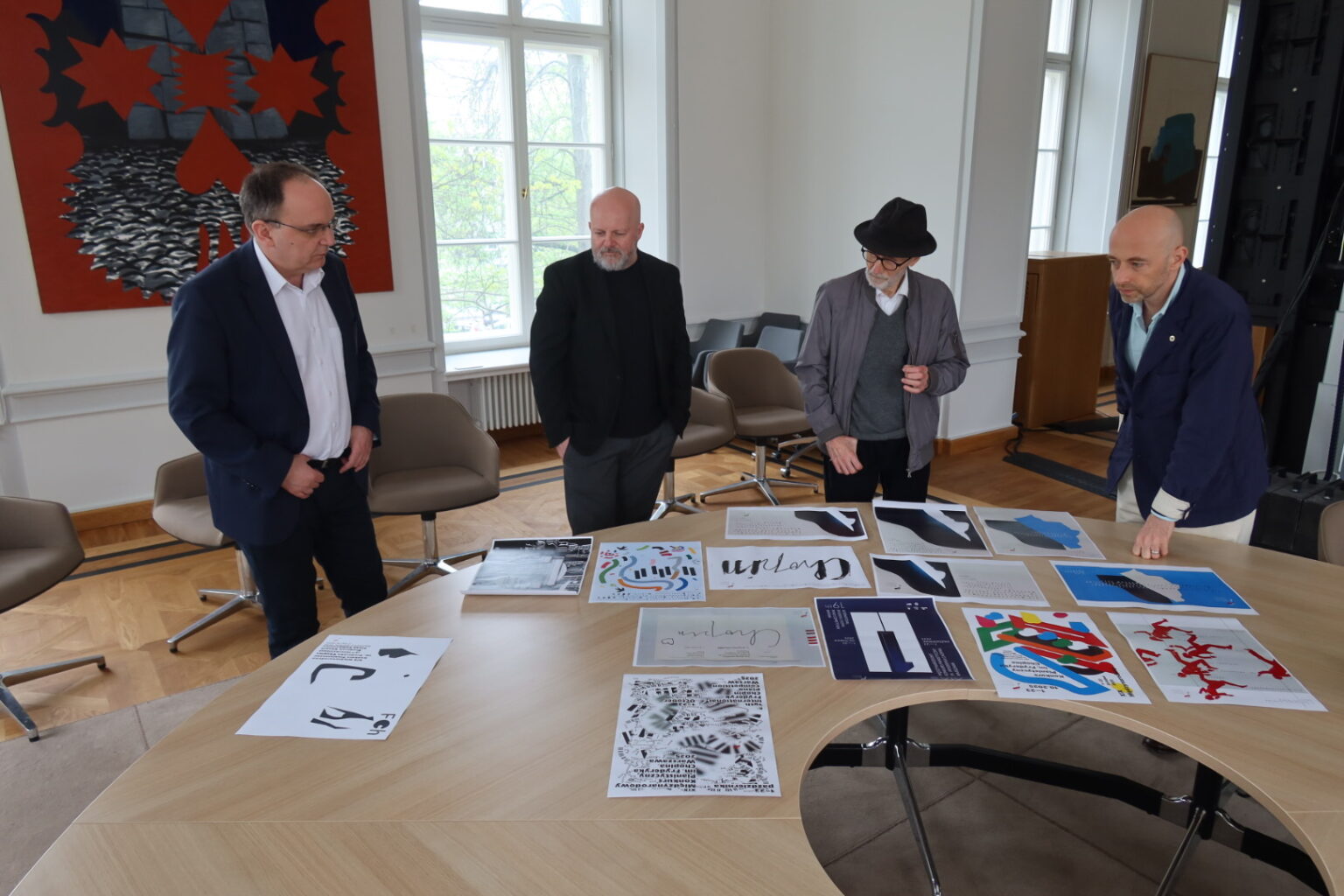 Czterech mężczyzn stojących przy okrągłym stole. Przed nimi rozłożone prace konkursowe.