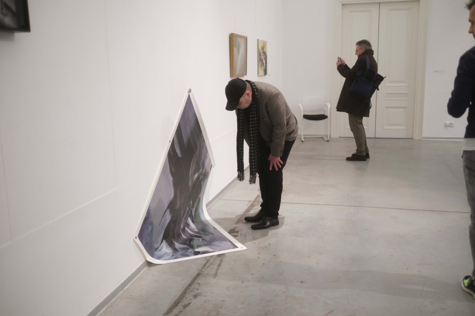 Schylony mężczyzna wygląda jakby chciał podnieść plakat, który został powieszony na ścianie galerii tak jakby się osuwał na podłogę