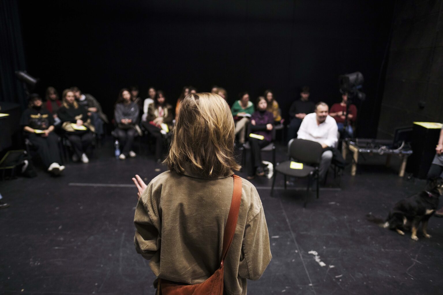kobieta odwrócona tyłem na scenie, mówi do ludzi siedzących przed nią na krzesłach