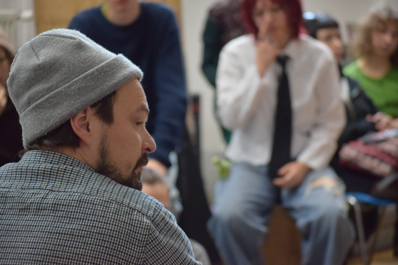 Zdjęcie od tyłu - profil mężczyzny w szarej czapce, na przeciwko niego siedzą osoby