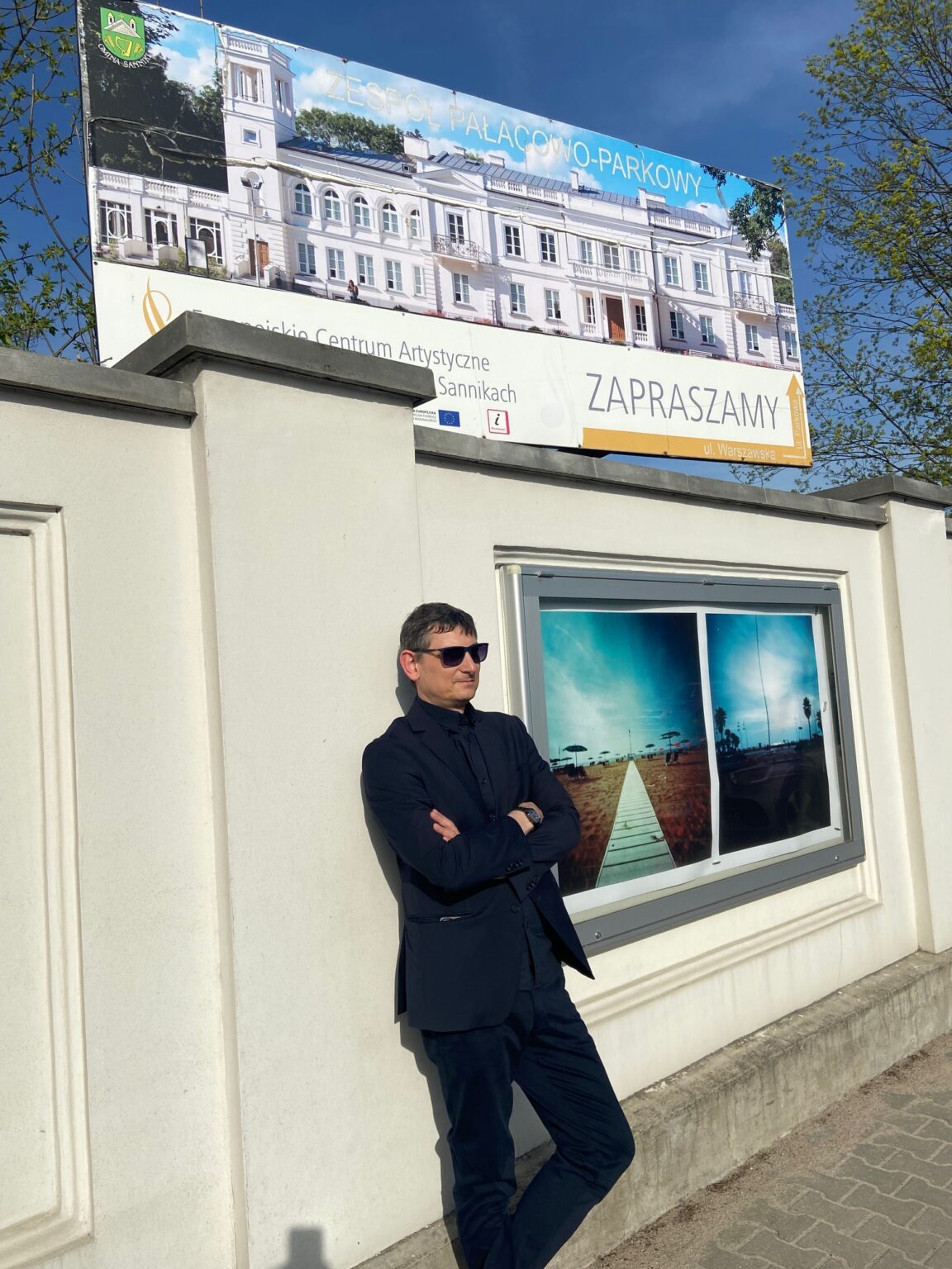 Mężczyzna w czarnym garniturze i okularach przeciwsłonecznych stoi obok gabloty, w której prezentowane są zdjęcia